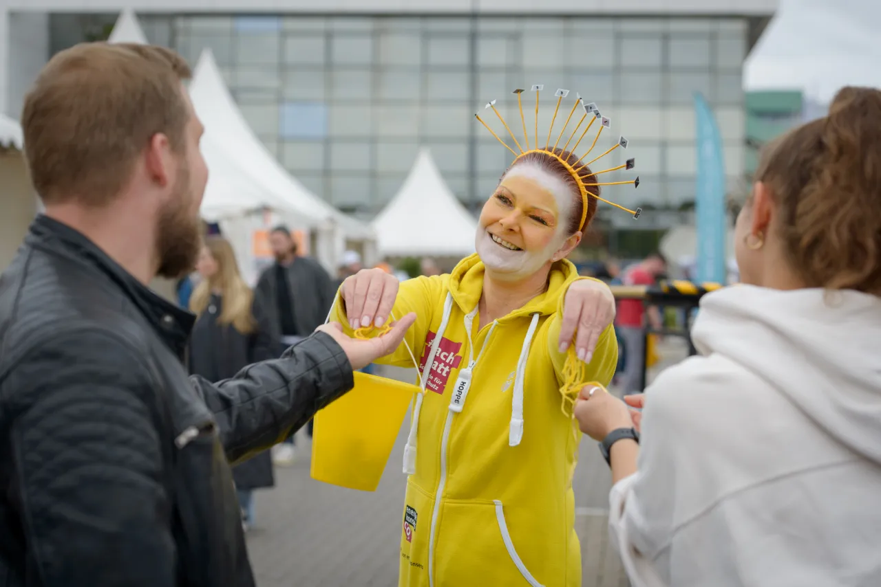 Maskottchen Sunna &uuml;bergibt Sonnen-Pf&auml;den an zwei Besucher*innen